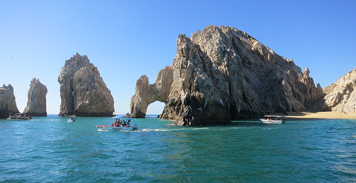 a group of people on a rock near the ocean with Arch of Cabo San Lucas in the background