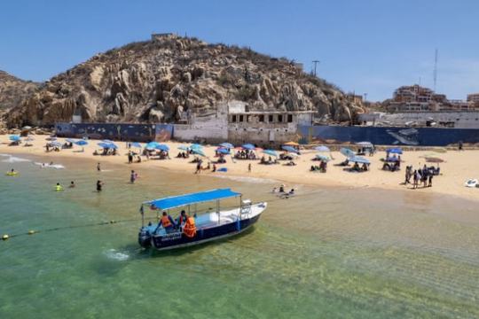 Boat near sandy beach with people, umbrellas, rocky hill, and buildings in background.