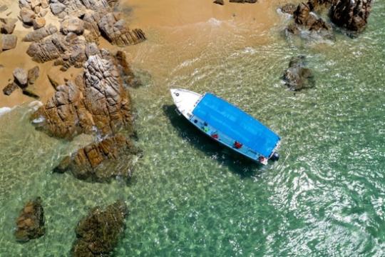 Aerial view of a boat with a blue tarp near rocky shore and clear green water.