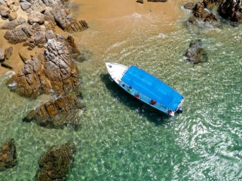 Aerial view of a boat with a blue tarp near rocky shore and clear green water.