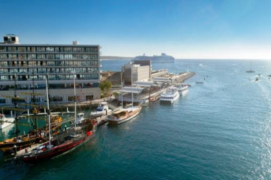 Harbor with docked boats, a waterfront building, and a distant cruise ship under clear blue sky.