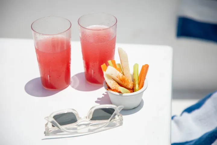 Two red drinks, a bowl of vegetable sticks, and sunglasses on a white surface.