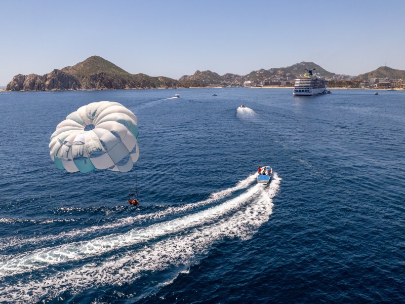 Parasailing over the ocean near a boat, with mountains and a cruise ship in the background.