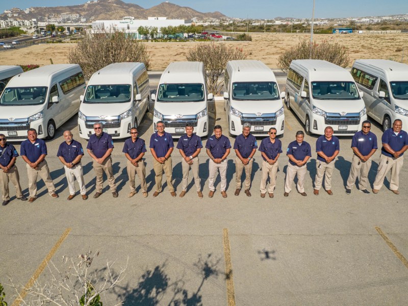 A row of 13 men in matching uniforms stand in front of parked white vans in a lot.