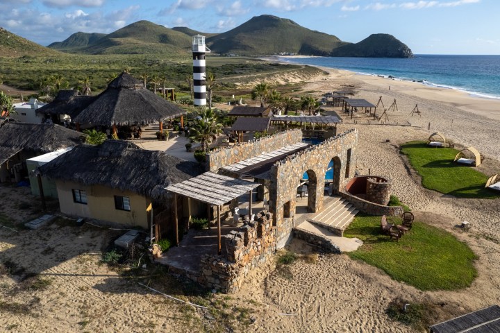 Coastal resort with straw huts, stone arches, and a lighthouse, set against mountains and ocean.