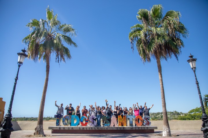 Group of people posing with raised arms by colorful Todos Santos sign under palm trees and clear sky.