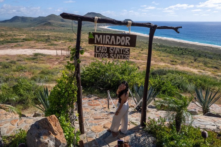 A woman poses under a mirador sign with ocean and mountains in the background.
