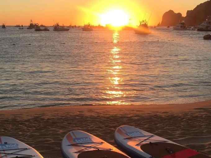 Sunset over ocean with paddleboards on sandy beach and boats in distance.