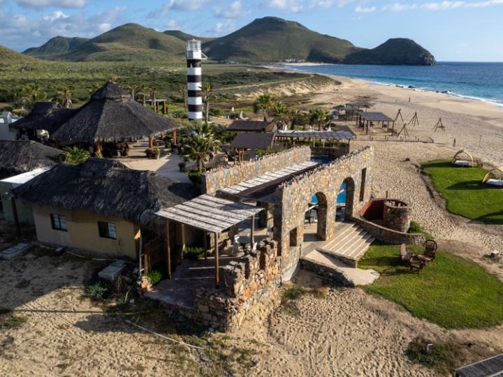 Beachfront resort with stone structures, thatched roofs, and a striped lighthouse overlooking the sea.