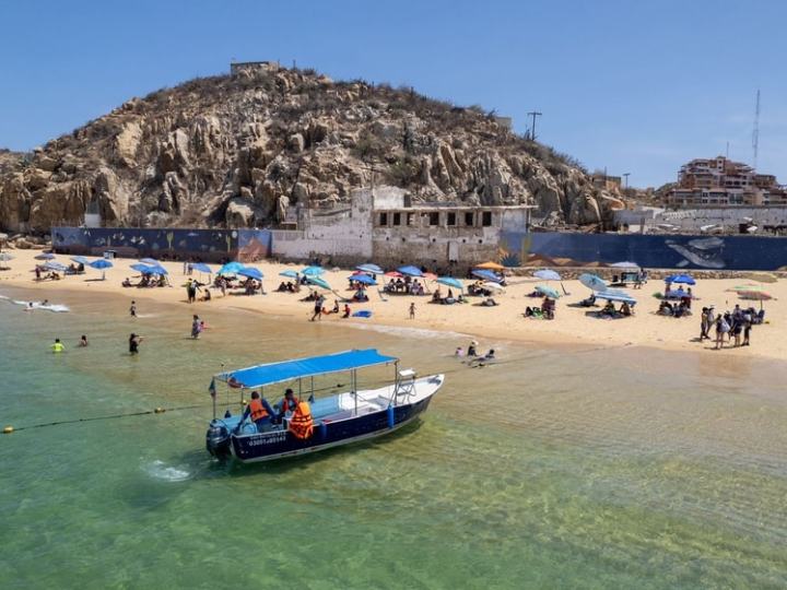 Beach with people, umbrellas, a motorboat on water, and rocky hill and buildings in the background.