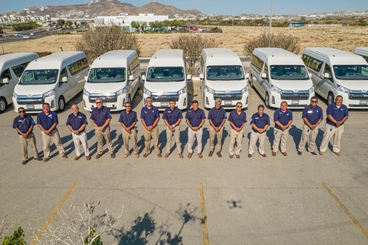 Eleven men in blue shirts stand in front of a row of white vans in a parking lot.