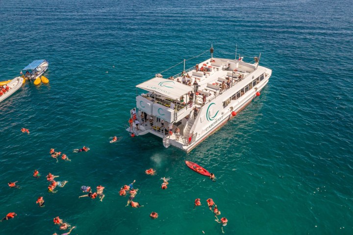 Aerial view of people swimming near a large boat and a small boat on clear blue water.
