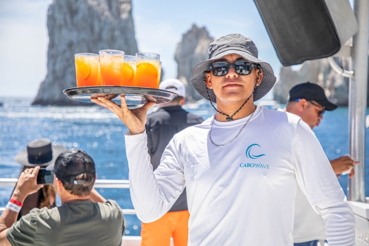 Server in hat holding tray with drinks on boat, ocean and rocks in background.